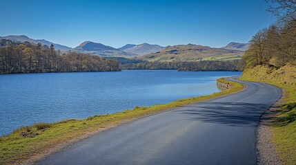 A beautiful road by the lake with mountains in the distance on a sunny day. The scenery is stunning, with the blue lake and green mountains contrasting against the black road.