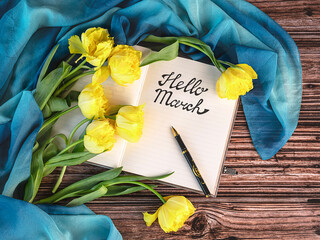 Hello March concept. An open notebook with the inscription hello March, a pen, a bouquet of yellow tulips, a blue shawl on a dark background.