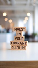 Four wooden blocks stacked on a white surface read "INVEST IN YOUR COMPANY CULTURE" in sans-serif font. A blurred bright background highlights the clean message about business and company culture.
