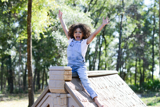 A joyful child in overalls sits on a playhouse roof with arms ra