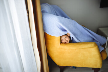 A delighted child pokes their head out of a blanket fort made with a chair, the cozy hideout