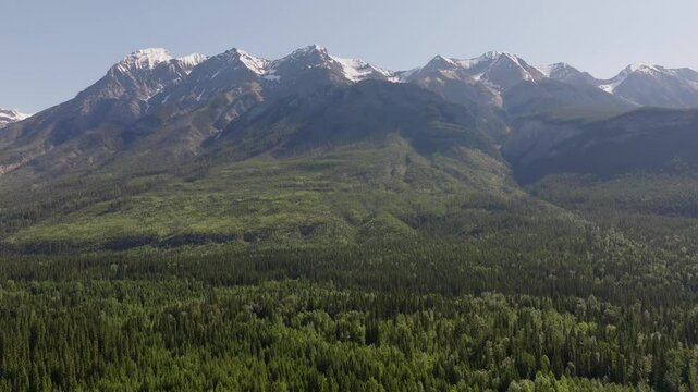 Drone pulling back from tall snowy mountains and cloudy sky and tilting downward showing vast pine forest wilderness near Yoho and Banff National Park in Cananda