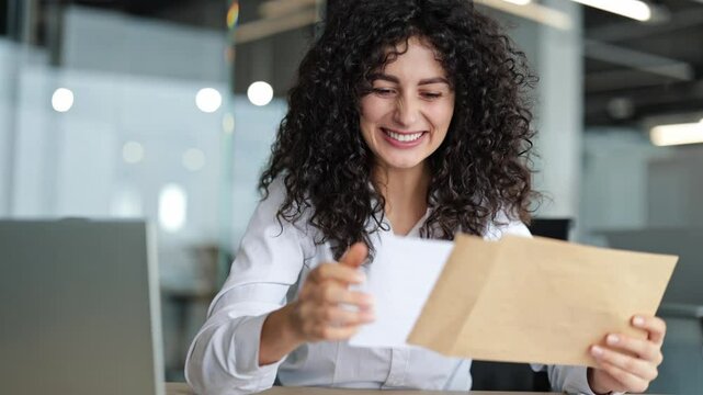 Excited businesswoman reading good news in a letter at her office desk. Curly-haired woman receives an unexpected job offer or notice of success.