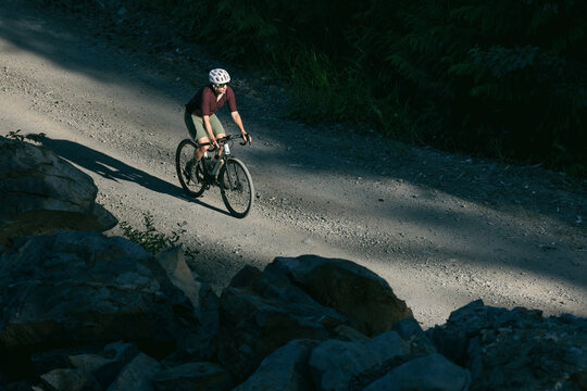 Cyclist on a gravel road winding through a dense forest with shadows.
