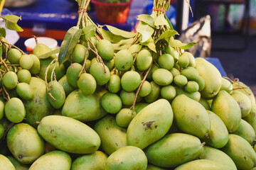 vibrant pile of green mangoes, both ripe and unripe, with some still attached to branches with leaves. The scene appears to be at a market or a fruit stand.