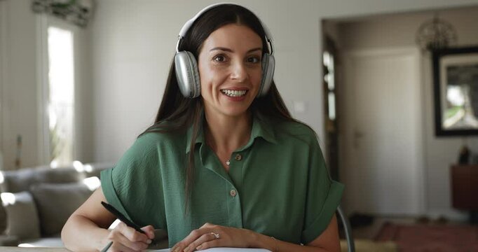 Beautiful woman wear headphones talk to teacher looking at camera, learning, gaining new knowledge, improve language skills, writes in copybook, make notes or assignment e-learning at home using tech