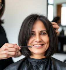 A cheerful woman with dark hair smiles while a hairstylist trims her bob haircut using a comb and scissors in a modern salon setting, creating a professional and welcoming atmosphere.