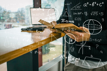 A person holding an open book surrounded by overlaid mathematical formulas, with laptop on wooden table in background. Education, science, and technology in modern setting