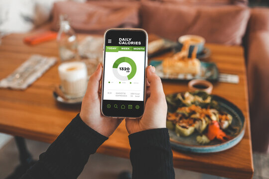 Hands holding smartphone displaying daily calorie tracker app with nutritional information. Table with plates of food and a drink is blurred in background.