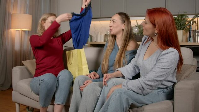 Cheerful charming mature woman taking out purchase from shopping bag, showing new nightie to lovely middle aged female friends , talking and sharing about quality and style at home.