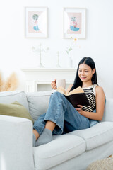 Vertical image of beautiful Asian woman lean on sofa hold a cup of  tea also enjoy to read the book with happiness in living room with day light.