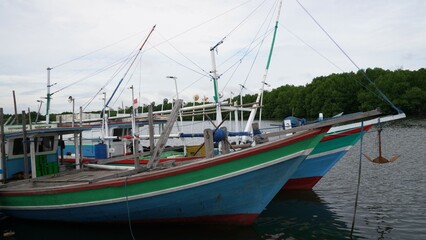colorful boats docked at a pier with a blue and white striped barrier and covered walkway. The serene waterfront scene includes reflections of the boats and pier in the calm water, set against a backd