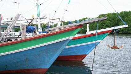 colorful boats docked at a pier with a blue and white striped barrier and covered walkway. The serene waterfront scene includes reflections of the boats and pier in the calm water, set against a backd