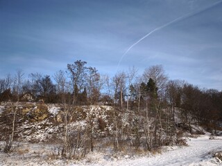 Winter landscape featuring a snow covered hillside with bare trees, a rock face, and a contrail...
