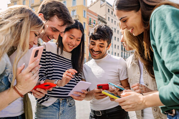 Happy students enjoying time together outdoors using mobile phones, sharing content and smiling....