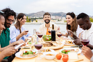 Happy group of multiracial young friends having dinner at rooftop summer party. Multiracial people laughing and having fun while drinking and eating sitting on table during celebration