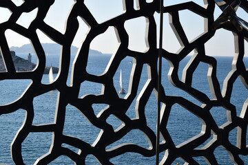 A sailboat glides across the sea, framed by the intricate modern architecture of the MUCEM in Marseille. A stunning juxtaposition of innovation and natural beauty.