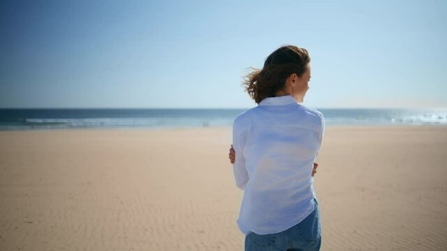 Back woman walking beach in white shirt windy day. Young brunette looking ocean