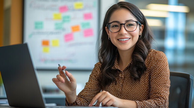 a photograph of a smiling, dark-haired woman with glasses, wearing a brown polka-dot blouse, sitting at a desk in a modern office. she's holding a pen and gesturing while looking at a laptop screen. a