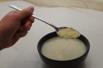 a man's hand showing the sleeve of a sweater, holding a spoon full of soup over a black bowl with noodle soup on a marble table
