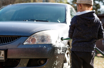 A little boy washes a car outdoors. A happy healthy little child washes the car with water from a hose, splashing merrily.