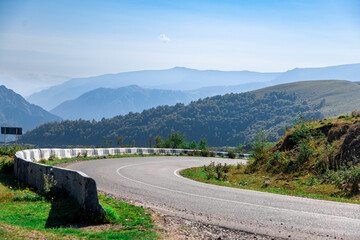 A mountain range with a winding road that is surrounded by trees. The road is curvy and has a white line painted on it. The mountains are covered in trees and the sky is blue