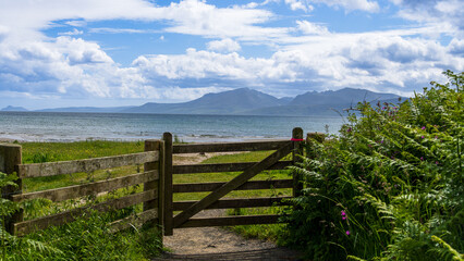 Scotland, Isle of Bute. Photo taken on: 08-06-2024. A serene coastal scene with a wooden gate, greenery, and mountains in the background.