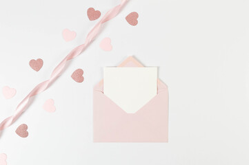 Top view of pink envelope, white card and pink ribbon on white background. Valentine's day, pink hearts, glitter hearts, love. Shiny rolled fabric, silk and satin material. Copy space, flat lay, post.