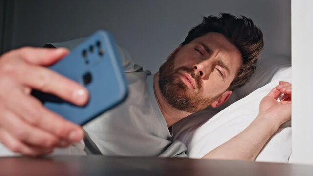Sleepy guy reaching smartphone in bed at sunny morning. Brunette man waking up