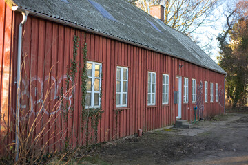 Old red wooden building with white windows and overgrown ivy located in a rural area during late afternoon light
