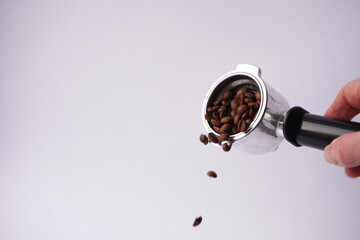 A close-up of a hand holding a portafilter filled with roasted coffee beans, with some beans...