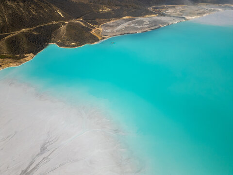 A turquoise tailings pond near a cement factory, surrounded by rugged hills. The vivid water contrasts with the barren land, creating a striking yet industrial scene of environmental impact