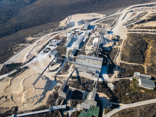 A large industrial cement plant surrounded by barren hills and dirt roads. Conveyor belts and...