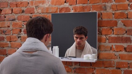 Young man in the morning stands in front of the mirror in bathroom with towel over neck, doing...