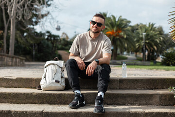 Handsome fashion hipster traveler man with trendy sunglasses in t-shirt with sneakers in backpack sitting and relaxing on steps in park in Barcelona