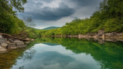 Obraz premium Serene river scene surrounded by lush greenery and rocky banks under a moody sky, reflecting the tranquil water surface.