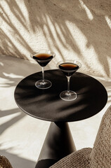 Black round table with two espresso martinis in glasses on the top, minimalist setting, shadow play, natural light, beige aesthetic, wicker armchairs, white concrete floor