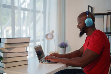 Focused black student wearing headphones, sitting at a desk and typing on a laptop, studying online with books nearby, showcasing modern education and self-improvement