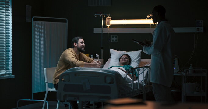 Woman patient rests in bed, accompanied by a man, with an African doctor holding medical records nearby.