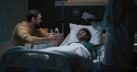 Girl is lying in a hospital room with a man seated beside her. Standing nearby is a doctor holding...