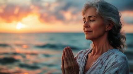 A senior woman practicing yoga on an ocean beach at sunrise. Her hands clasped together in a prayer-like position.