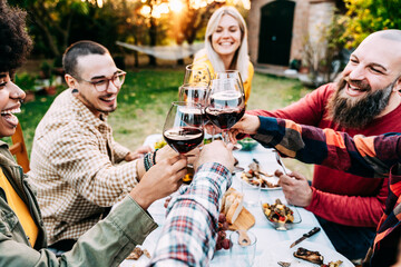 Multiracial friends having fun at barbecue dinner party in garden restaurant - Millennial people cheering red wine sitting at outside bar table at summer - Youth lifestyle, food and beverage concept