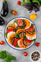 Eggplant caprese salad with balsamic sauce and fresh basil on a plate on a gray concrete background. Vegetarian food. Top view, copy space.