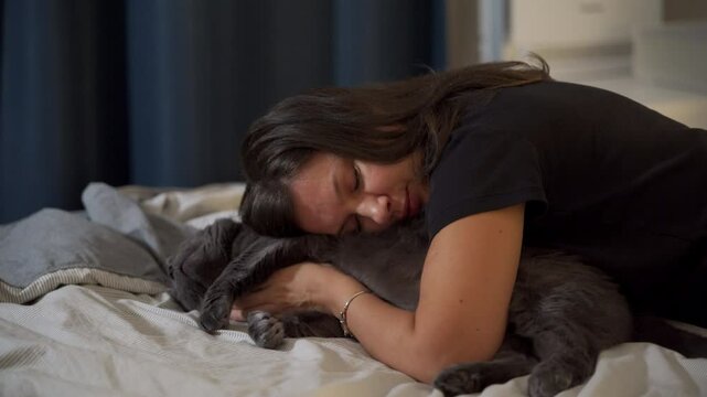 Adult 44 year old woman and gray domestic cat lying on bed in bedroom. Happy woman hugging and kissing her cat, stroking his belly.