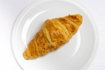 A freshly baked golden croissant on a white plate, placed on a white wooden table, showcasing its flaky and buttery texture as a perfect breakfast or snack option.