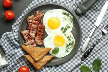 Tasty fried eggs, bacon and pieces of toast served on black table, flat lay