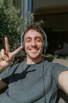 Young man takes selfie video call on couch outdoors. Smiling guy shows peace sign with phone in hands. Relaxed atmosphere. Sunny day. Casual wear. Modern tech. Lifestyle photo. Indoor outdoor space.