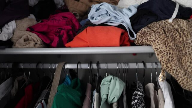 A messy closet shelf filled with a variety of folded and unfolded clothes in different colors and patterns. The disorganized setup highlights the need for decluttering and organization.