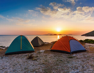 Three colorful camping tents are set up along the rocky Aegean coastline during a spectacular sunset with golden sun reflecting on the calm Mediterranean waters.