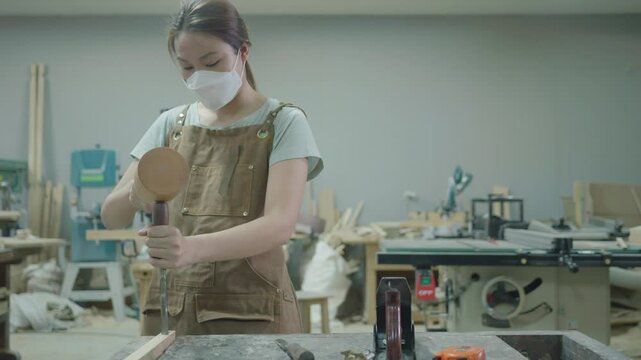 A young woman is focused on her woodworking project in a studio, showcasing craftsmanship and creativity. Wearing protective gear, she skillfully uses a tool amidst a workspace filled with wood materi